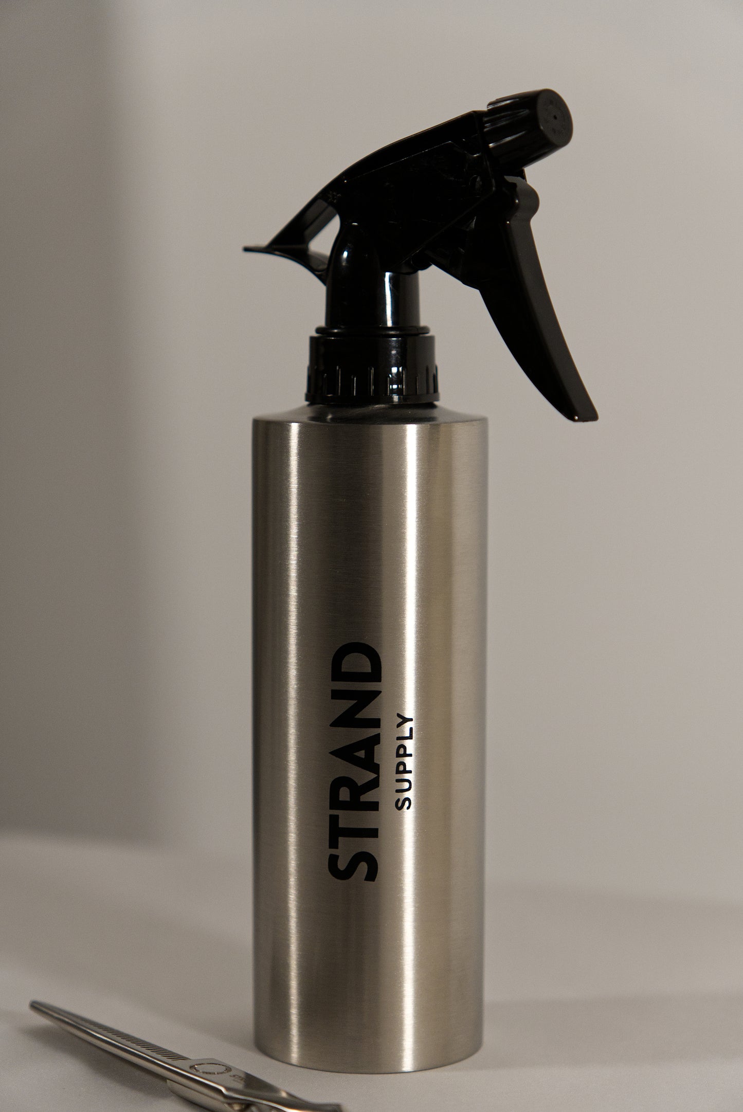 Silver spray bottle with black nozzle labeled 'Strand Supply' on a neutral background